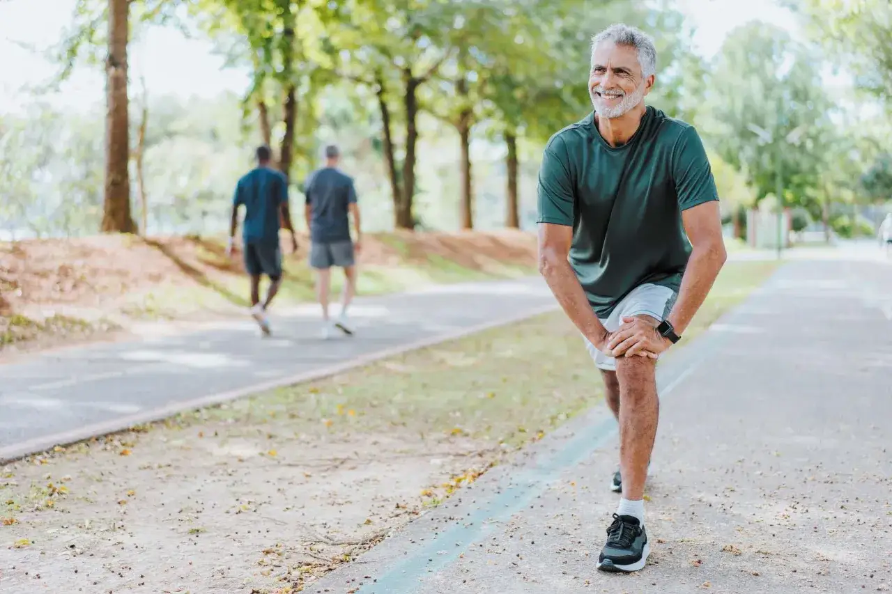 Ein &auml;lterer Mann dehnt sich l&auml;chelnd auf einem Weg im Park. Er beweist, dass ist spazieren gehen sport. Im Hintergrund gehen zwei M&auml;nner spazieren.