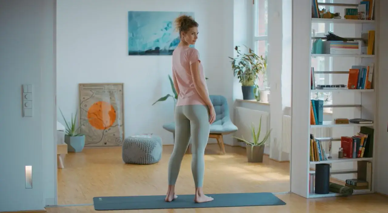 Frau in Sportkleidung auf Yogamatte, bereit f&uuml;r &Uuml;bungen. Ein Regal voller B&uuml;cher und Pflanzen im Hintergrund. Vielleicht hilft dies bei der Bew&auml;ltigung des BWS-Syndroms.